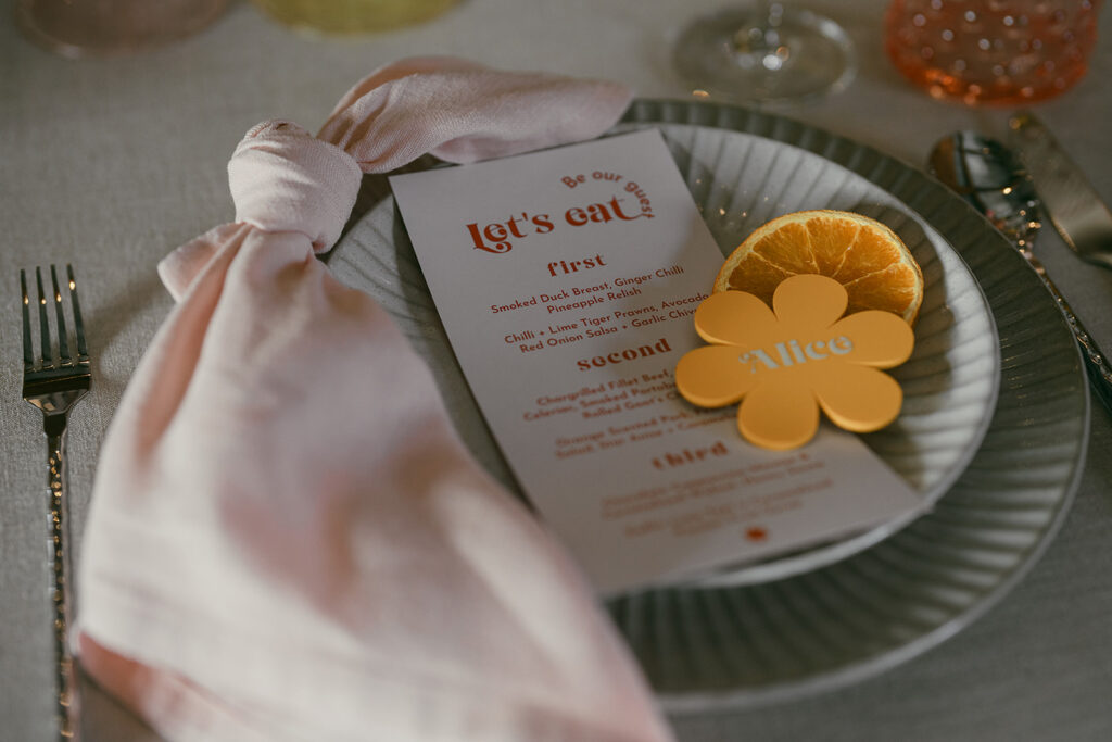 Place setting with menu in the middle of the starter plate, pink napkin knotted and placed on the left side, dehydrated slick of orange and a flower shaped retro place name.