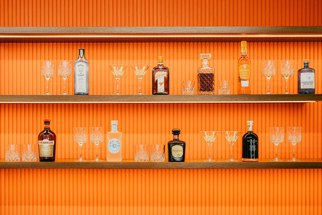 Bright orange bar backdrop lined with various crystal glasses in pairs amongst spirits such as gin, whiskey, contreau and espresso martini mix.