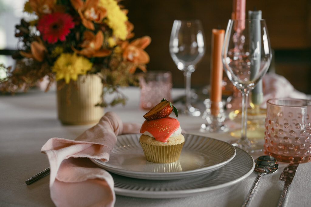 Closeup of the pink-frosted cupcake with a half strawberry on top. The cupcake is sitting in the middle of the starter plate with the glassware, napkin, florals and candles blurred in the background