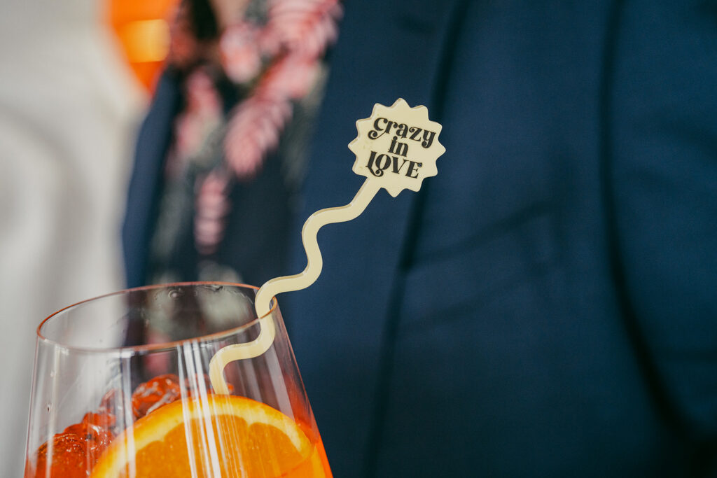 Close up shot of the swizzle stick that reads "crazy in love" in the glass of aperol spritz. Held by the groom and you can see pops of pink colour from his shirt blurred in the background