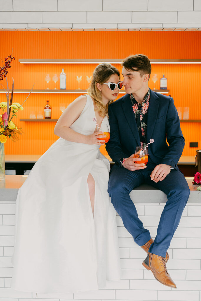 Bride and groom sitting on the bar with the orange back wall in background. She has white sunglasses on and they are holding Aperol Spritz each