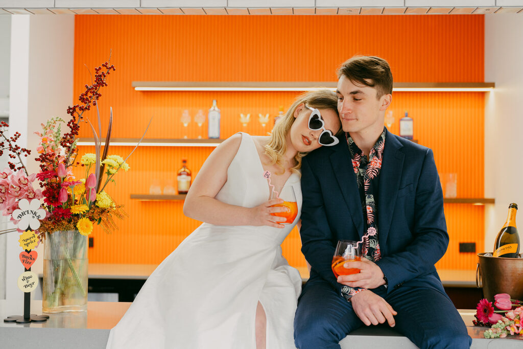 Bride and groom sitting on the bar in front of the bright orange bar backdrop. Bride is wearing heart sunglasses and has her head on the grooms shoulder. 