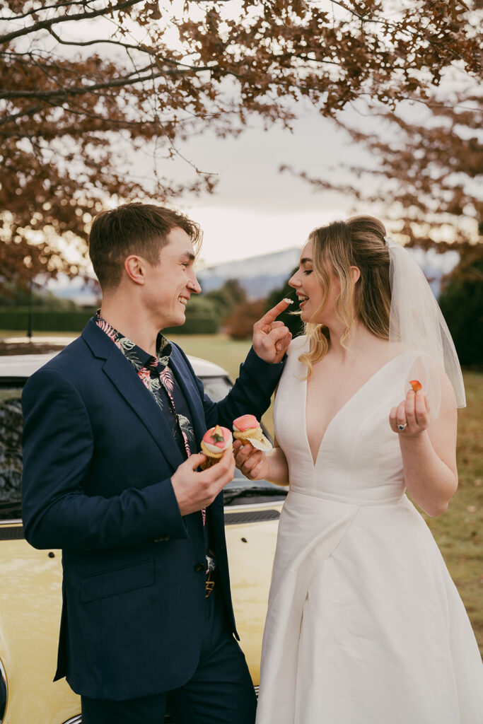 Bride licking cupcake frosting of the groom's finger in front of a yellow mini in the afternoon light