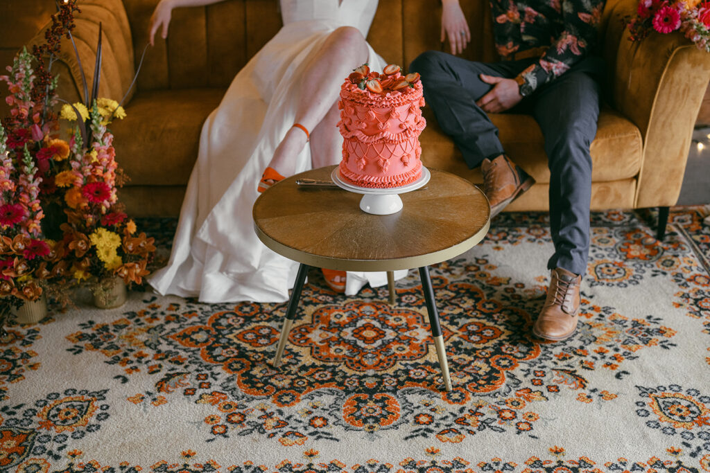 Bride and Groom sitting on the gold retro couch with a retro round coffee table in front of them with the bright pink retro iced cake - ready to cut.