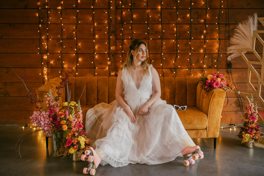 Bride sitting on the gold retro couch in roller skates and a floaty, chiffon gown. She is surrounded by colourful retro florals and a backdrop of fairy lights. The colour of the image is moody orange and gives sunset vibes.
