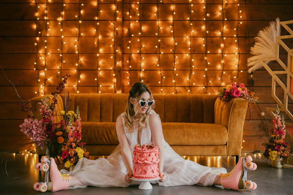 The bride is sitting on the floor in pink roller skates with a bright pink retro cake in front of her. She has white heart sunglasses on and is looking coy and playful.