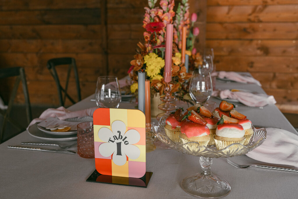 Full tablescape showing colourful retro florals in the background and retro table number and dish of pink frosted cupcakes in the foreground