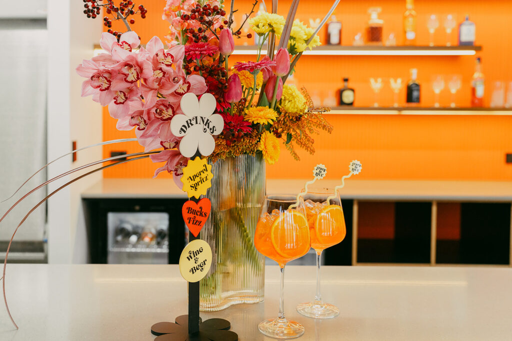 Bright bar set up with retro drinks signage in the shapes of circles, stars, flowers and hearts. 2 glasses of orange aperol spritz's with swizzle sticks that read "crazy in love" on them
