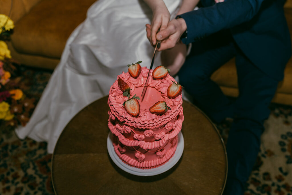 Close up of the bright pink retro iced cake with the knife about to slice into it. The cake is topped with half strawberries. The bride and groom are blurred in the background