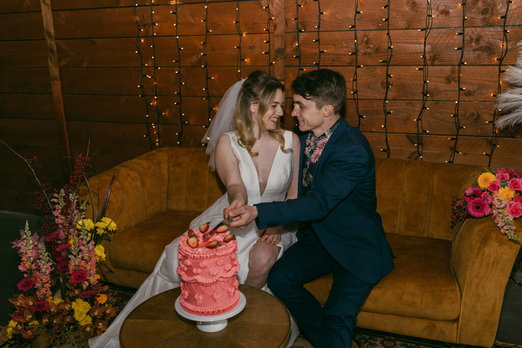 Bride and groom sitting on the gold retro couch, looking into each others eyes about to cut a retro looking cake with bright pink icing.