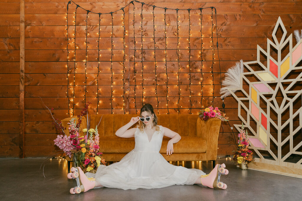 The bride is sitting on the floor in front of a fairy light backdrop and gold retro couch. She has pink roller skates on and  heart sunglasses. She looks fun and retro and ready to party