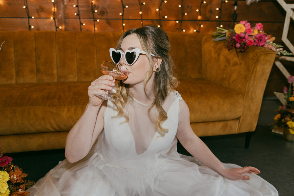 Close up of the bride in full, floaty chiffon dress. She has white sunglasses on and is taking a sip of champagne from a gold-rimmed, coupe glass. This image give fun, flirty, party vibes.