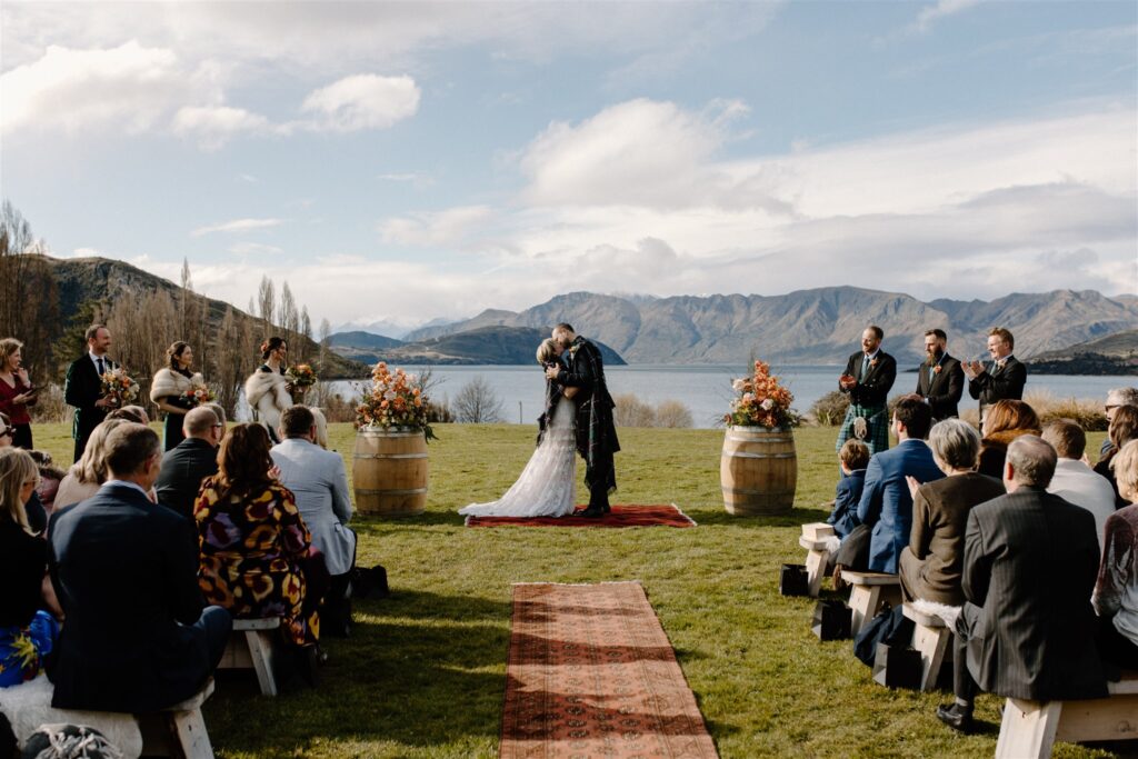 Outdoor wedding venue. Bride and groom kiss with their bridal party and guests looking on at their ceremony site. They stand on a bright red rug, between two wine barrels covered in florals, with the lake and mountains behind them.