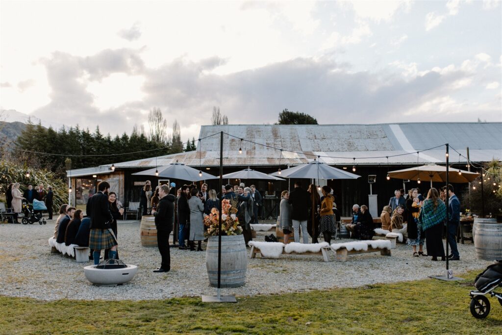 Outdoor wedding venue with guests standing around the outside of an old woolshed. Festoon lights, wine barrels and sheepskins add to the ambiance of the setting.