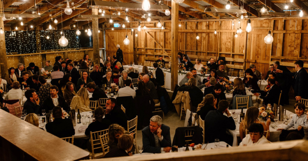 Wedding venue of woolshed converted into an intimate wedding reception. Big festoon lighting hanging from the ceiling and guests sitting at large, square tables.