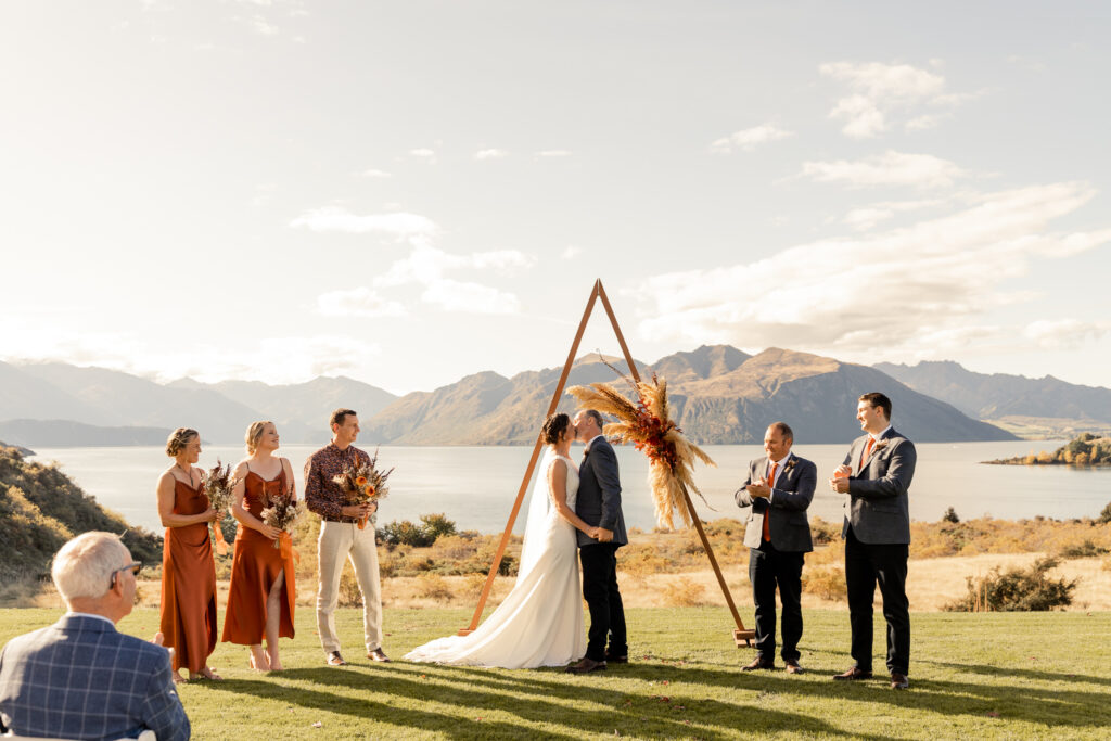 Bride and groom kiss in front of triangle wedding arch with Lake Wanaka in the background. 