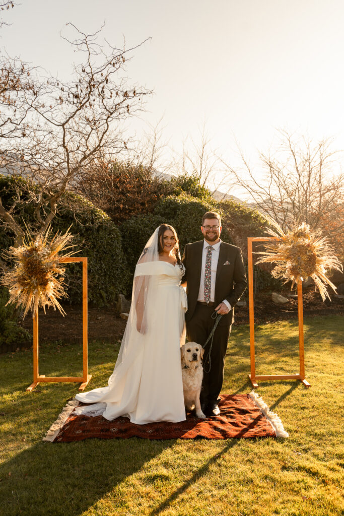 Outside wedding venue with bride and groom on a colourful rug on the grass. Their dog is standing between them.